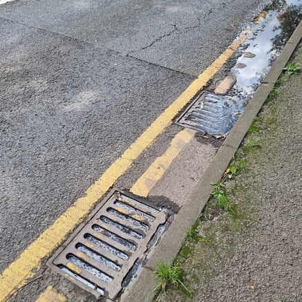 Road drain full of silt, causing a build up of water, oil and road waste